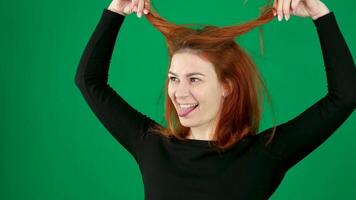 el niña sonrisas, endereza su cabello, muestra su lengua, en un verde fondo, un niña con rojo pelo en negro ropa. video