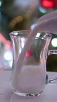 gegen das Hintergrund von ein Weihnachten Baum Milch gegossen in ein transparent Glas das Kamera bewegt sich um Gießen heiß Milch in ein Glas Tasse video