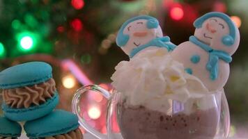 un taza de cacao con crema dos merengue muñecos de nieve macarrones en el antecedentes de un Navidad árbol Navidad celebracion vaso vaso hermosa sabroso atmosférico antecedentes sitio para publicidad video