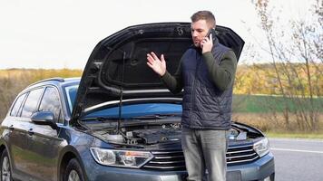 triste hombre en pie hablar en teléfono cerca el roto coche abrió el capucha la carretera ayuda reparar estrés problema emergencia seguro auto lento movimiento. transporte, de viaje concepto video