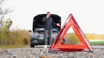 Road side warning triangle, warning oncoming traffic of a broken down car, with a man using his cell phone to call for assistance. Transportation, traveling concept video