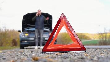 la carretera lado advertencia triángulo, advertencia venidero tráfico de un roto abajo auto, con un hombre utilizando su célula teléfono a llamada para asistencia video