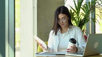 jung charmant weiblich Freiberufler mit Laptop Computer zum Entfernung Job während Sitzung im modern Kaffee Geschäft Innere mit Unterlagen video