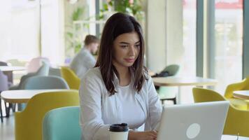 giovane affascinante femmina libero professionista utilizzando il computer portatile computer per distanza lavoro mentre seduta nel moderno caffè negozio interno. bellissimo caucasico donna Lavorando su netbook durante mattina prima colazione nel bar bar video