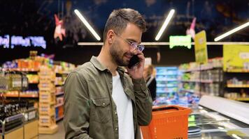 jovem homem com compras cestas dentro uma supermercado faz compras, escolhe produtos e fala em a telefone, consulta com dele esposa sobre a compras Lista video