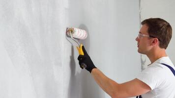 Side view of happy young male painter in blue overall and gloves painting wall, with roller in grey color while working on interior design of apartment video