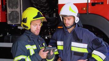 Two firemen in full equipped clothes standing outside at the fire truck with a tablet in hands and deciding what to do for extinguishing fire video