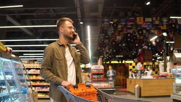 Young man with shopping trolely in a supermarket talks on the phone, consults with his wife about the shopping list video