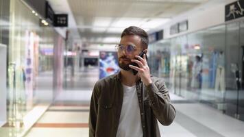 bonito jovem homem falando em uma Móvel telefone e sorridente enquanto caminhando dentro Shopping video