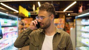 Young man walks in a supermarket makes purchases, chooses products and talks on the phone, consults with his wife about the shopping list video