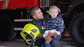 dapper brandweerman in uniform Holding weinig opgeslagen jongen tegen de achtergrond van een brand motor video