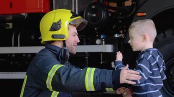 dapper brandweerman in uniform knuffels een weinig jongen met een speelgoed- tegen een brand motor video