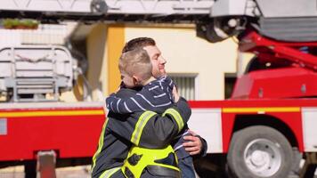 mutig Feuerwehrmann im Uniform Umarmungen ein wenig Junge mit ein Spielzeug gegen ein Feuer Motor video