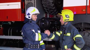 Two firefighters in protective clothing in helmets with fire engine, friendly handshake video