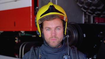 Close up portrait of strong serious fireman in helmet and full equipment standing next to car with flashing lights on and looking into camera video