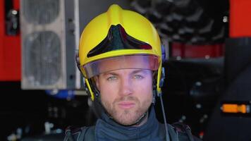 Close up portrait of strong serious fireman in helmet and full equipment standing next to car with flashing lights on and looking into camera video