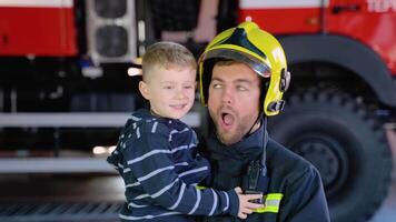 grappig brandweerman in uniform Holding weinig grappig jongen tegen de achtergrond van een brand motor video