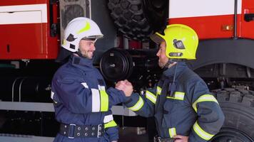 Two firefighters in protective clothing in helmets with fire engine, friendly handshake video