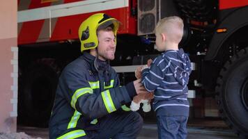 dapper brandweerman in uniform en weinig jongen met een speelgoed- tegen de achtergrond van een brand motor video