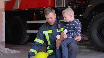 Brave firefighter in uniform holding little boy with a toy against the background of a fire engine video