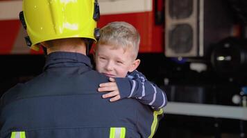 Brave firefighter in uniform holding little saved boy against the background of a fire engine video