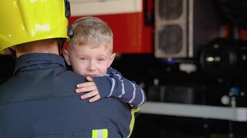 dapper brandweerman in uniform Holding weinig opgeslagen jongen tegen de achtergrond van een brand motor video