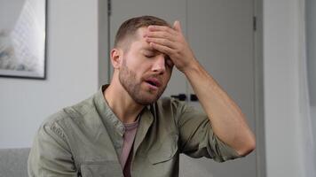 se sentir stressé. beau jeune homme frustré touchant sa tête et gardant les yeux fermés assis sur le canapé à la maison video