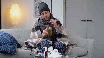 hälsa, influensa och människor begrepp - sjuk ung man i hatt insvept i en filt dricka varm te på Hem. man uppvärmningen hans händer. uppvärmning säsong video