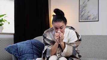 Young woman, sits on the couch at home under a plaid, sneezing in a napkin, feels weak, unwell, needs treatment and medicines. Sneezing, runny nose, cough video