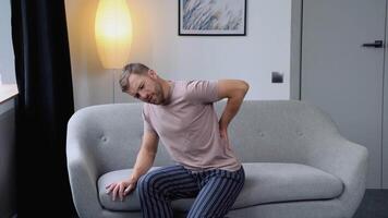 People, healthcare and problem concept - unhappy young man suffering from pain in back while sitting on sofa at home video