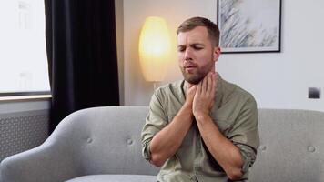 Terrible pain in his throat. Frustrated handsome young man touching his neck while sitting on the couch at home video