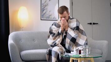 Sneezing, runny nose, cough. Unhealthy sad young man, sits on the couch at home under a plaid, sneezing in a napkin, feels weak, unwell, needs treatment and medicines video