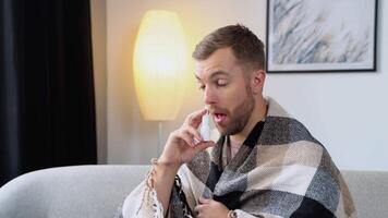 A young man wrapped in a blanket sprays a nasal spray while sitting on sofa video