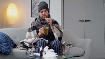 Health, flu and people concept - sick young man in hat wrapped in a blanket drinking hot tea at home. man warming his hands. Heating season video