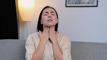 Frustrated young woman touching his neck while sitting on the couch at home. Terrible pain in his throat video