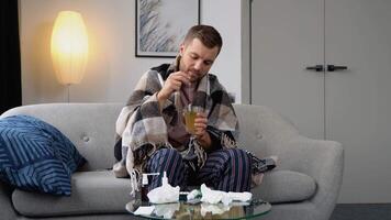 Health, flu and people concept - sick young man wrapped in a blanket drinking hot tea at home. man warming his hands. Heating season video