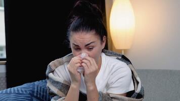 Young woman, sits on the couch at home under a plaid, sneezing in a napkin, feels weak, unwell, needs treatment and medicines. Sneezing, runny nose, cough video