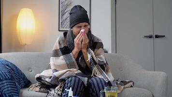 Sick frozen man sit on sofa, wear hat holds tissue blowing runny nose got fever, catch cold, heap of meds nearby. Seasonal influenza symptoms, flu, unheated flat concept video