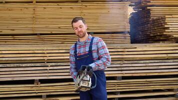 joven masculino trabajador con un motosierra en Tablas de madera depósito. concepto - rebaja de Tablas de madera video