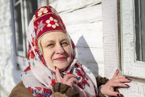 An elderly Russian woman in a traditional headdress, kokoshnik, poses against the backdrop of a village house. photo