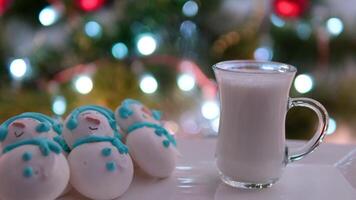 un vaso de caliente Leche cerca el Navidad árbol merengue muñecos de nieve en un blanco plato delicioso bocadillo cena gasto hora celebrando Navidad sano comida color ligero bombillas video