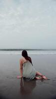 meisje in een jurk zittend Aan de zwart zand Aan de oceaan Aan een achtergrond van golven video