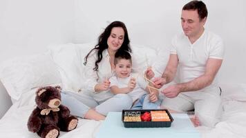 family eating sushi in bed at home ordering food delivery husband wife and little boy parents showing master class delicious appetizing sushi white clothes happy family life love at home video