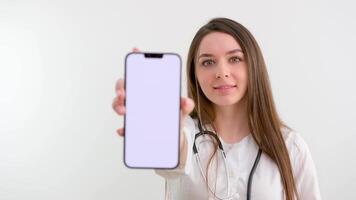 mujer médico o enfermero vistiendo un laboratorio Saco y estetoscopio señalando a un blanco móvil teléfono con un satisfecho sonrisa cerca arriba terminado un gris antecedentes video