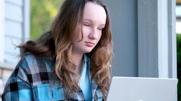 adolescent fille séance dans de face de portable en train de regarder film ou Faire devoirs en ligne en train d'étudier à la recherche pour emploi comme adolescent à la recherche vers l'avant à côté hausse les épaules soumissionner Jeune femme européen en plein air sur porche de maison video