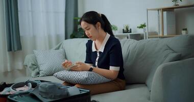 Side view of Asian teenager woman sitting on sofa making checklist of things to pack for travel, Preparation travel suitcase at home. photo