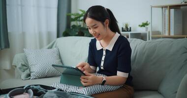 Portrait of Asian teenager woman sitting on sofa planning vacation trip and searching information on digital tablet, travel and lifestyle. photo