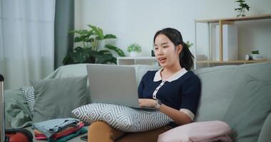 retrato de asiático adolescente mujer sentado en sofá utilizando ordenador portátil para preparar reserva hotel y avión boleto para viajar. mochilero viaje concepto. foto