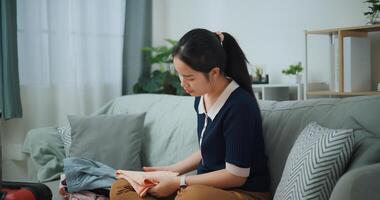 Side view of Asian teenager woman sitting on sofa packing travel luggage with clothes for traveling trip, Preparation travel suitcase at home. photo