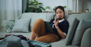 retrato de asiático adolescente mujer sentado en sofá participación crédito tarjeta haciendo en línea pago en digital tableta, preparación viaje maleta a hogar. foto